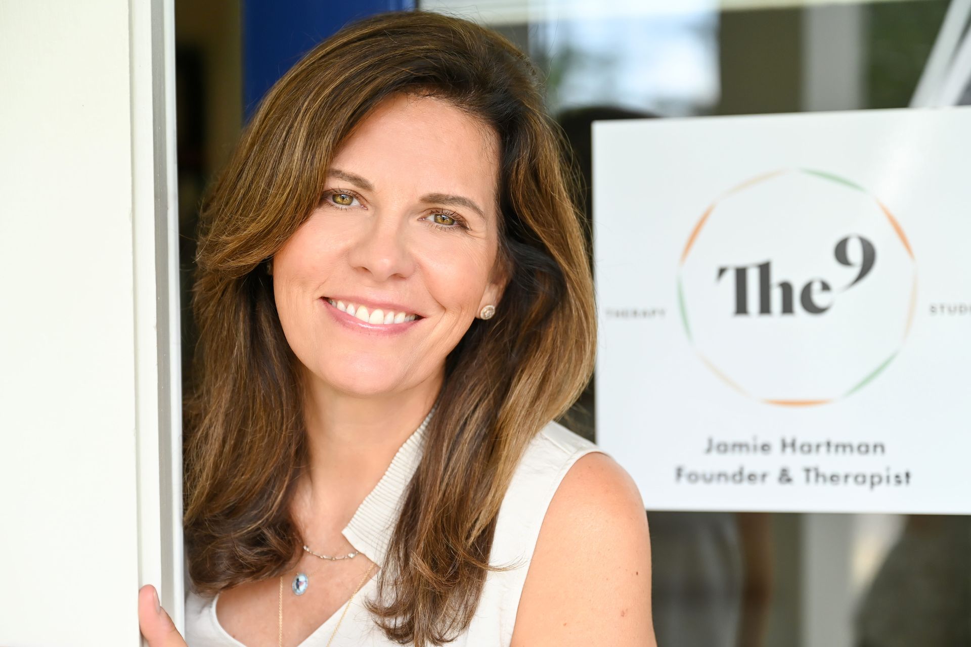 Smiling woman in white blouse beside a glass sign reading “The 9” and “Founder & Therapist”