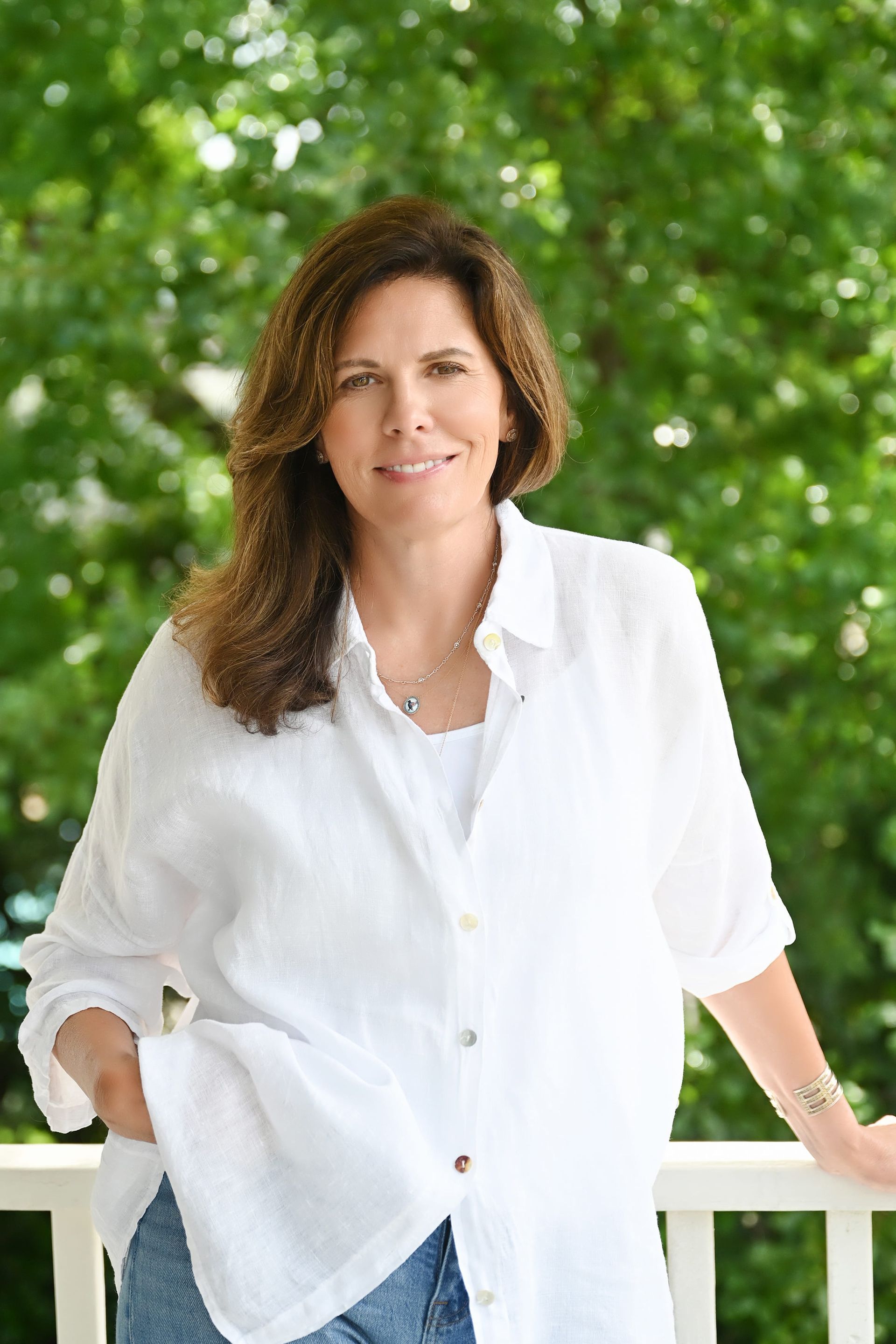 Woman in a white shirt standing outdoors by a white railing with green foliage behind her