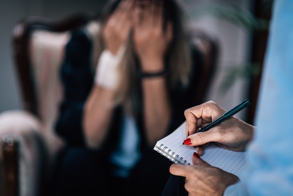Person writes notes while another seated person covers their face in distress.