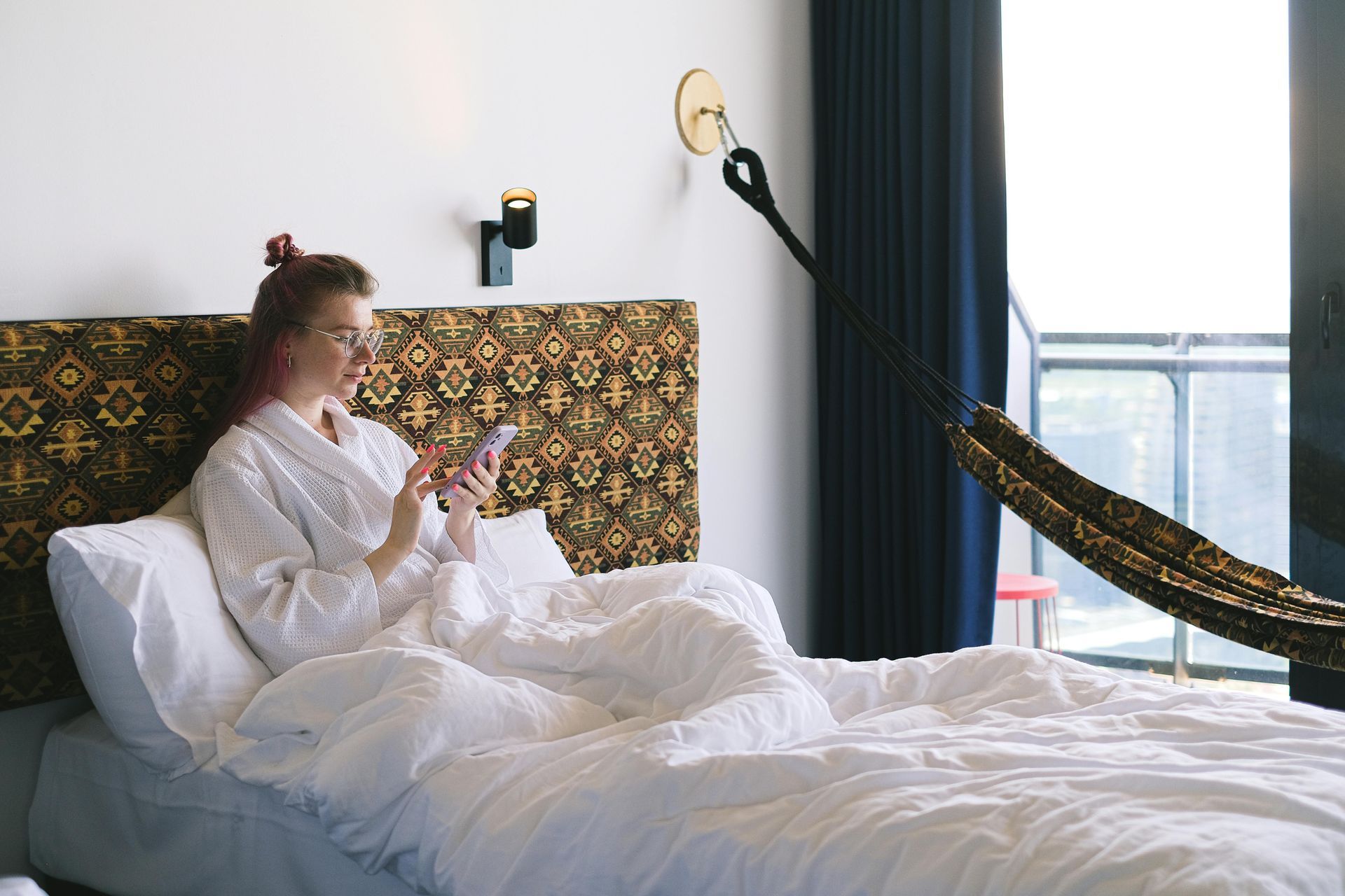 Person sitting on a bed in a bright bedroom, looking at a phone near a hammock chair and window.