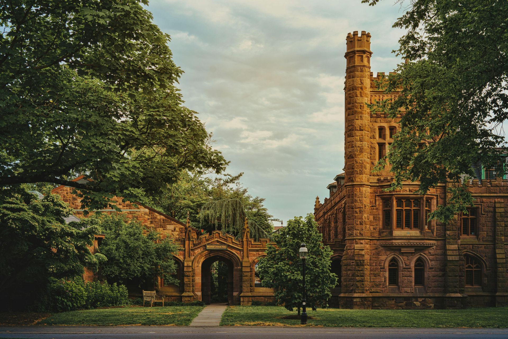 Historic brick building and arched entrance amid lush green trees under a cloudy sky