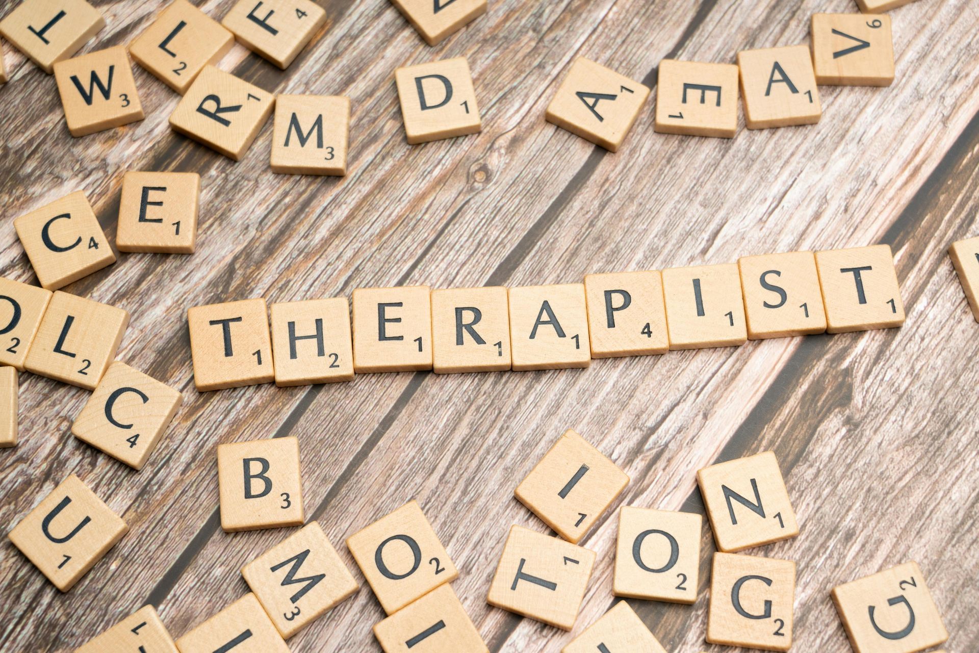 Scrabble tiles spelling “THERAPIST” on a wooden surface, surrounded by scattered letter tiles