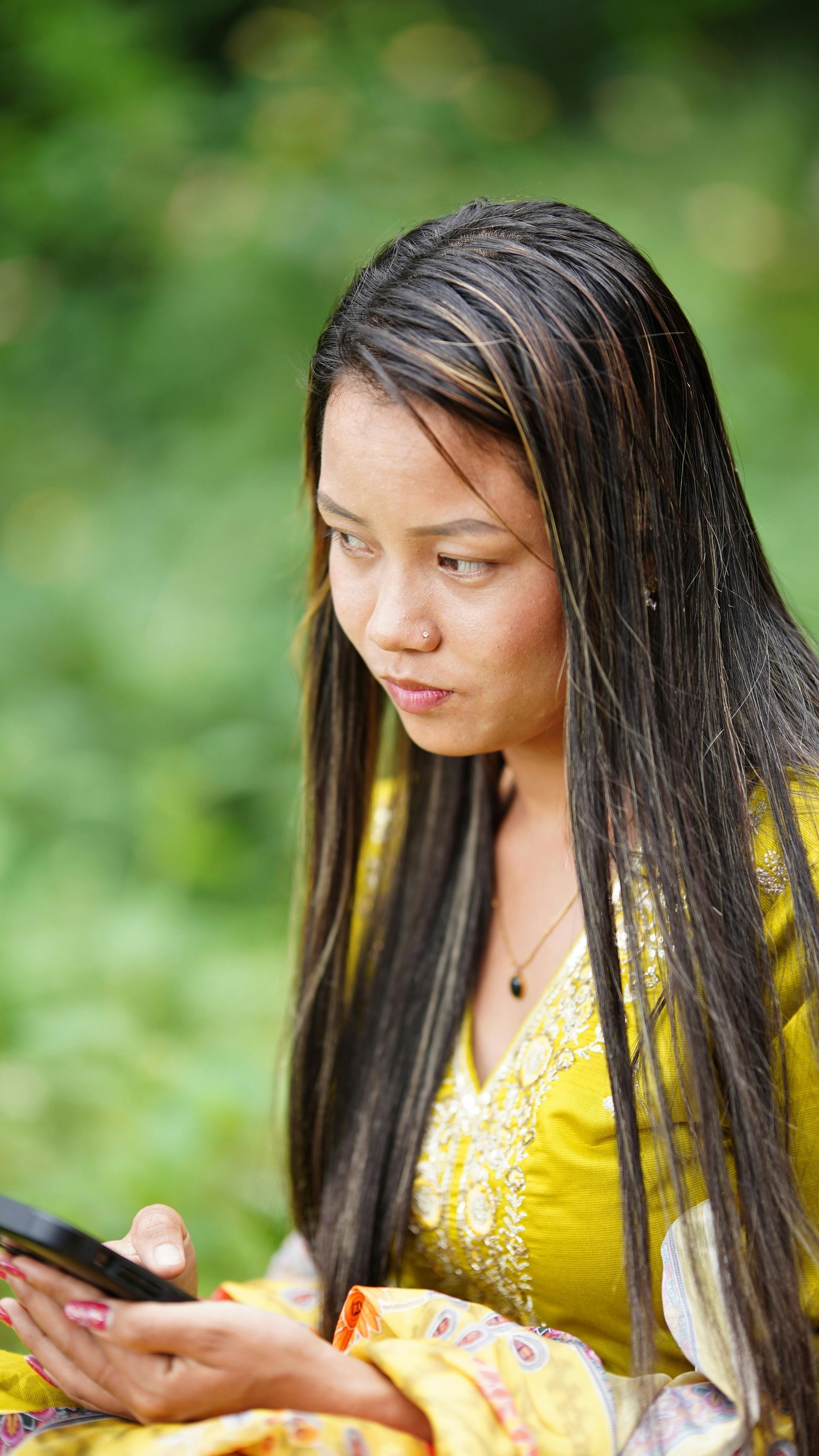 Woman in a yellow dress looking down at a phone outdoors with a blurred green background