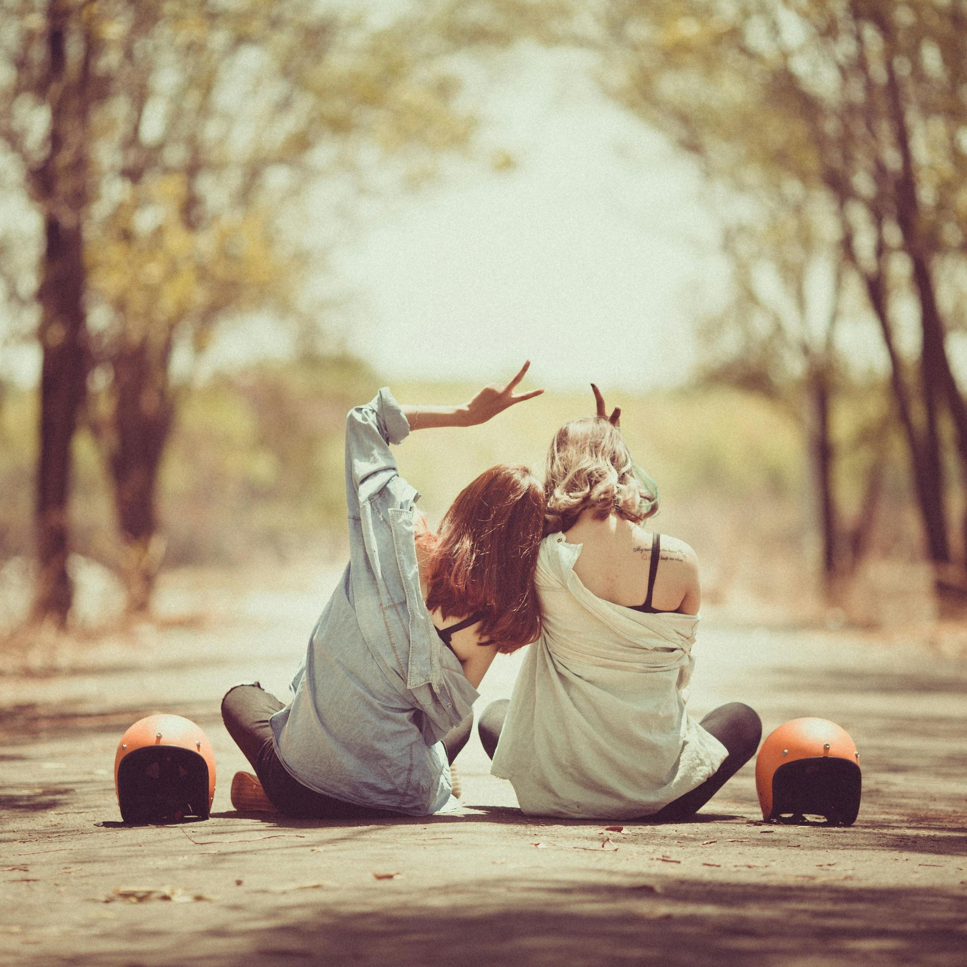 Two people sit on a road between orange helmets, raising peace signs in a sunny tree-lined tunnel