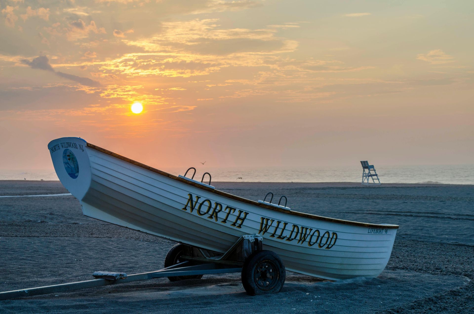 Boat named “North Windward” on a sandy beach at sunset, with a lighthouse in the distance.