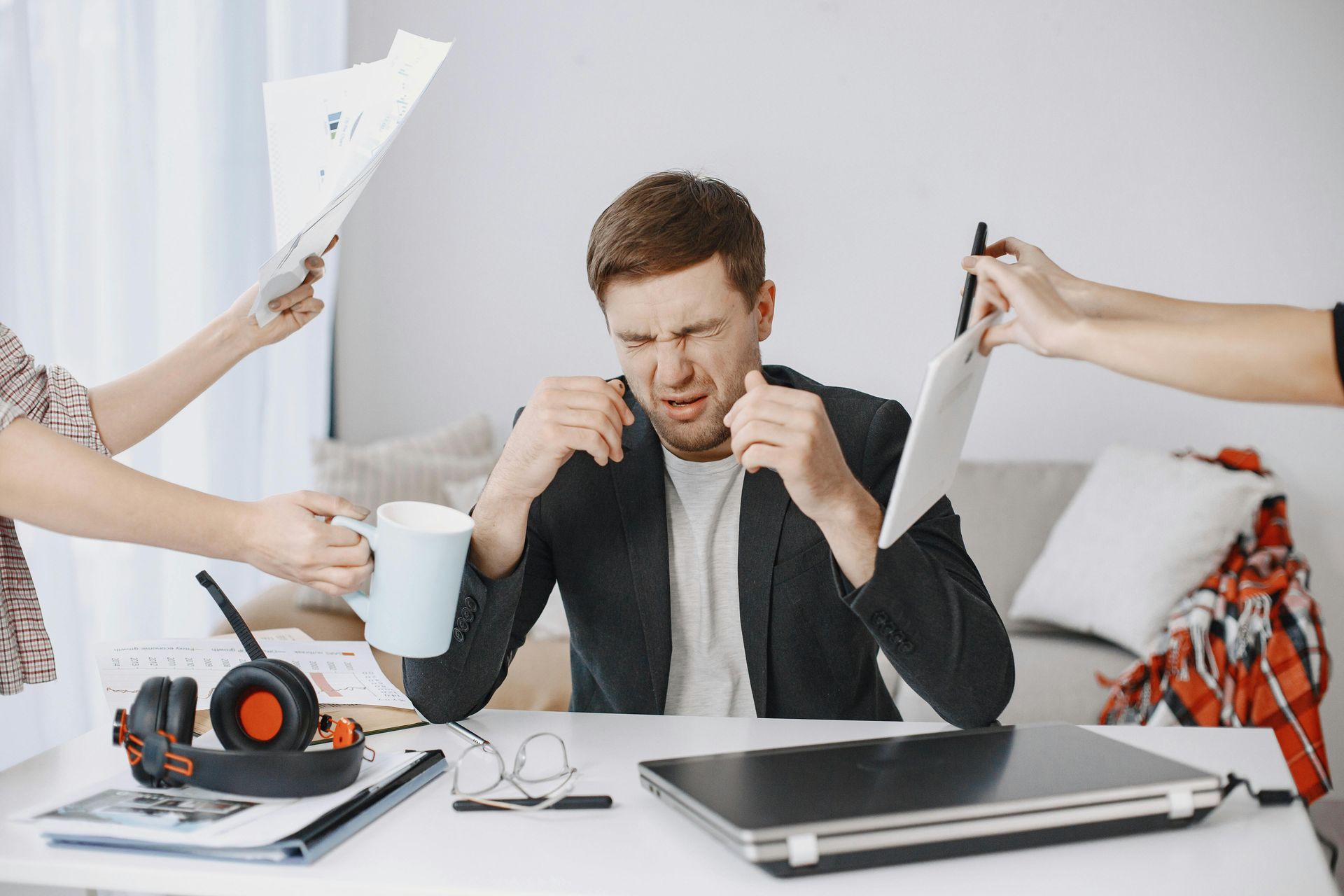 Man at desk looking stressed while coworkers’ hands extend papers and a cup toward him, with laptop and headphones nearby