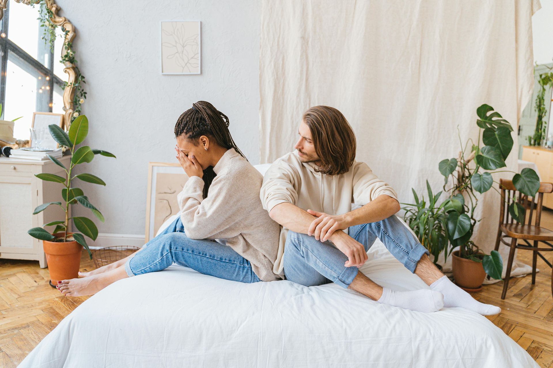 Two people sitting back-to-back on a bed in a bright room with plants and white curtains