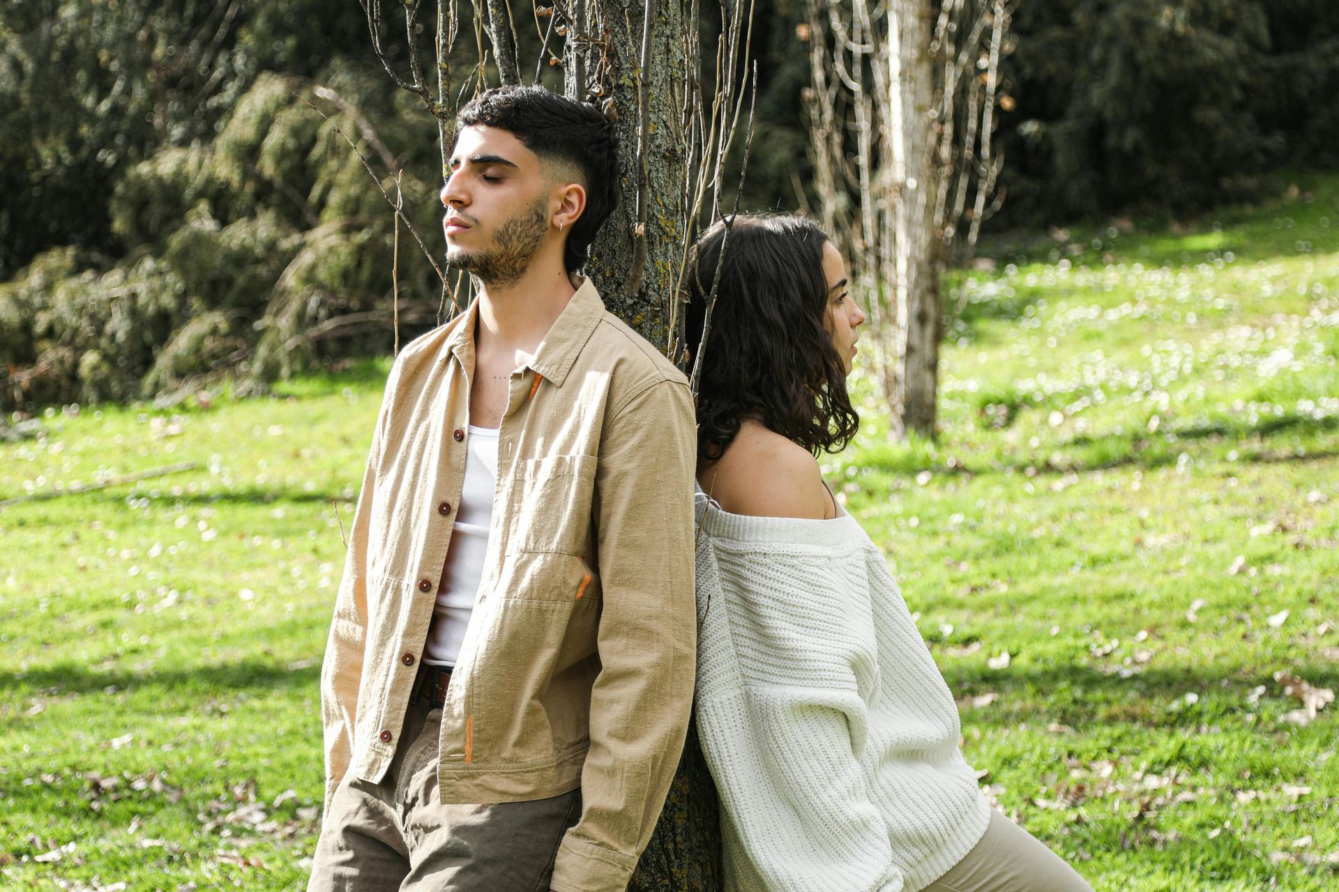 Two people back-to-back in a sunny grassy park, one in a tan jacket and the other in a white sweater.