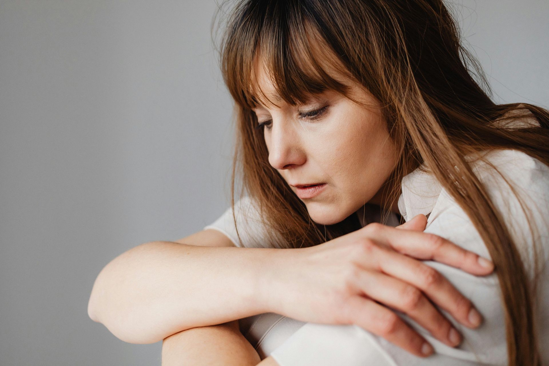 Woman in a white shirt sitting with arms wrapped around knees, looking down pensively against a gray wall