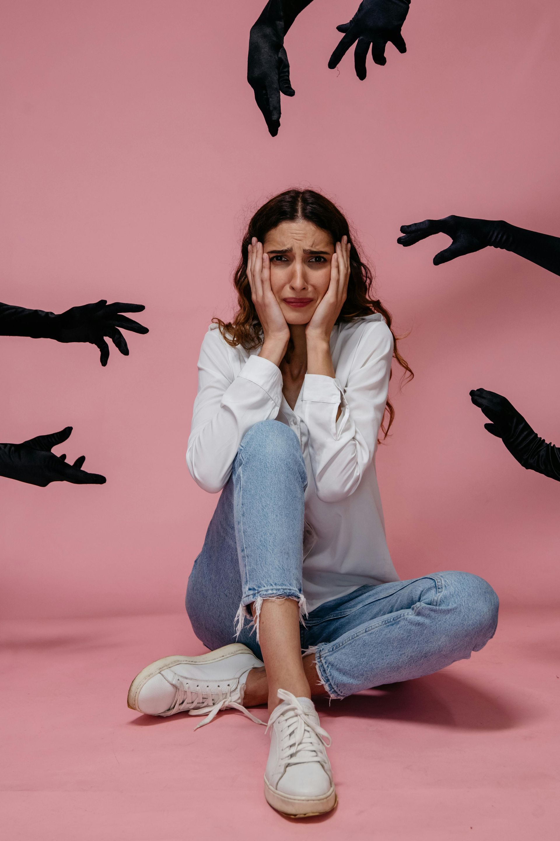 Woman sitting on a pink backdrop, holding her face, surrounded by reaching black hands