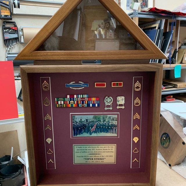 Framed military honor plaque with medals, insignia, and a group photo on display in a workshop.