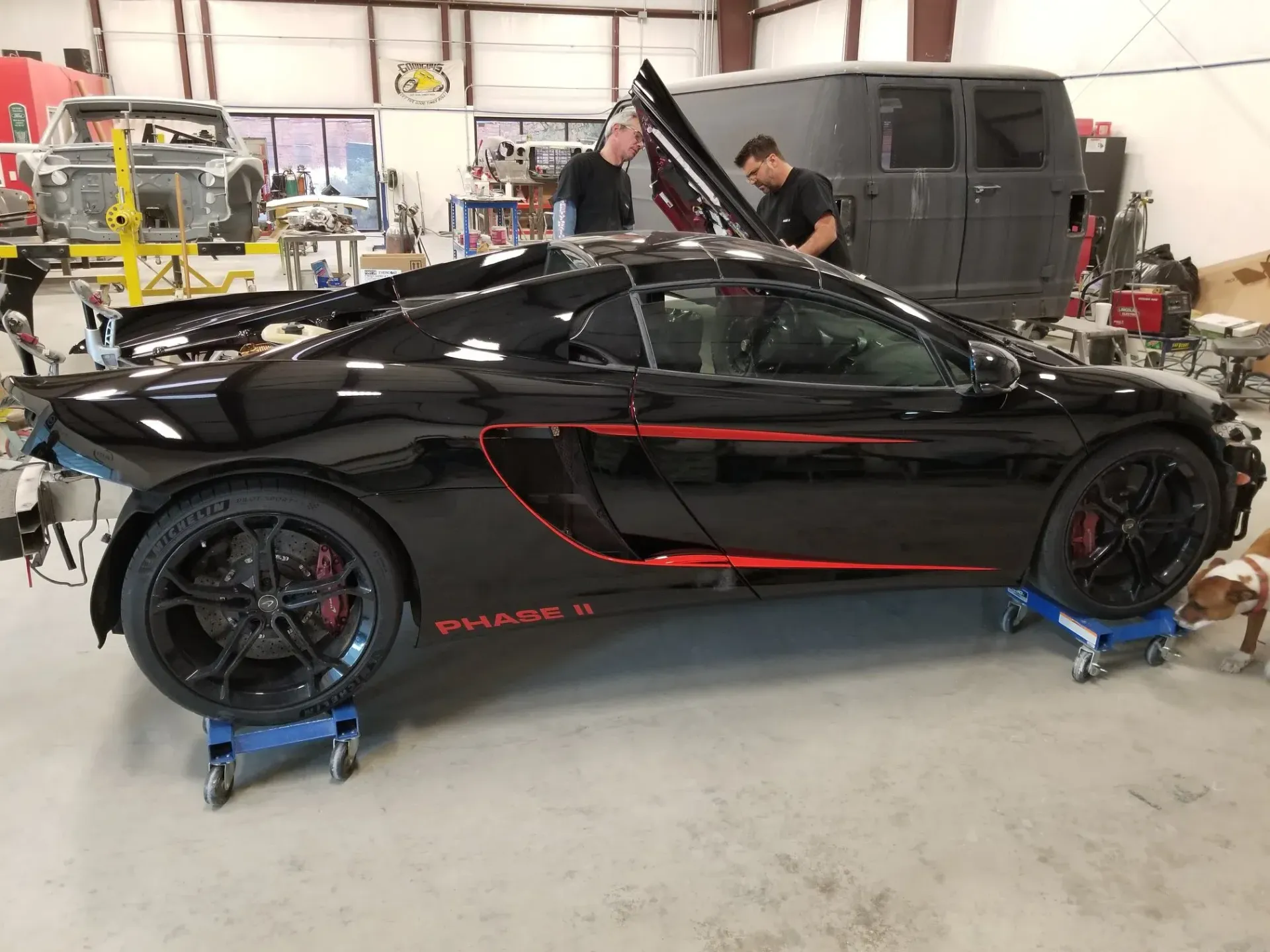 A black McLaren sports car with red accents is on dollies in a workshop while two mechanics work on the open engine bay.