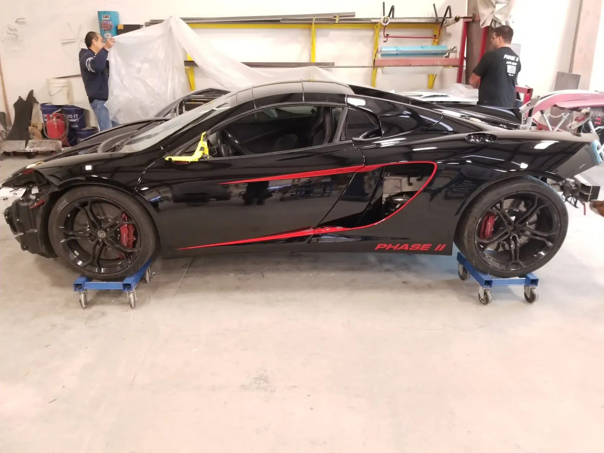 A sleek black sports car with red accents sits on automotive dollies in a workshop while two individuals work nearby.
