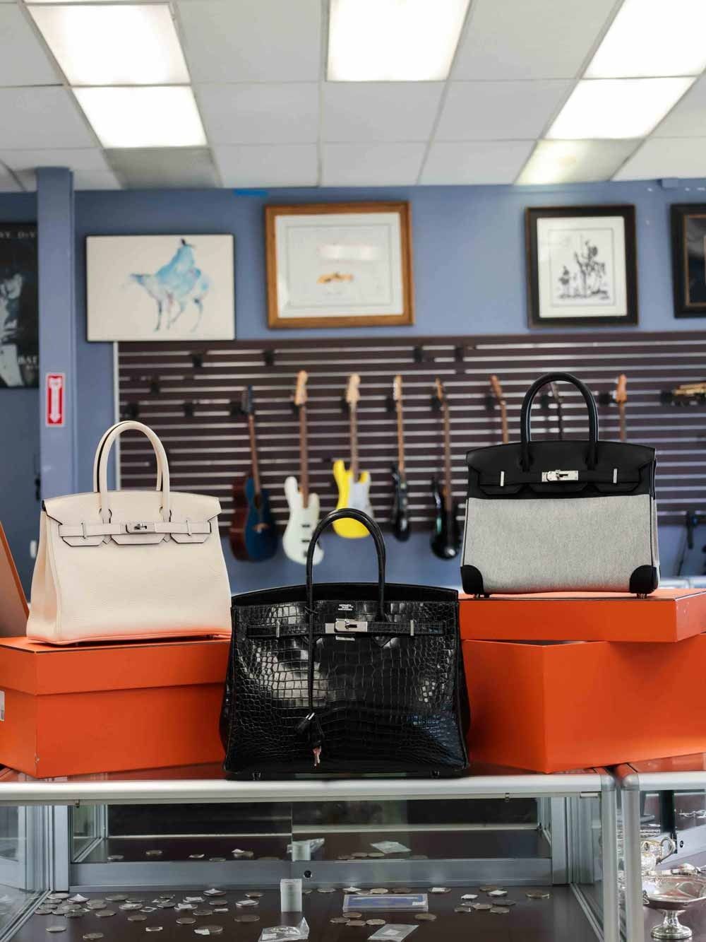 Handbags displayed on orange boxes atop a glass counter in a shop with framed art on the wall