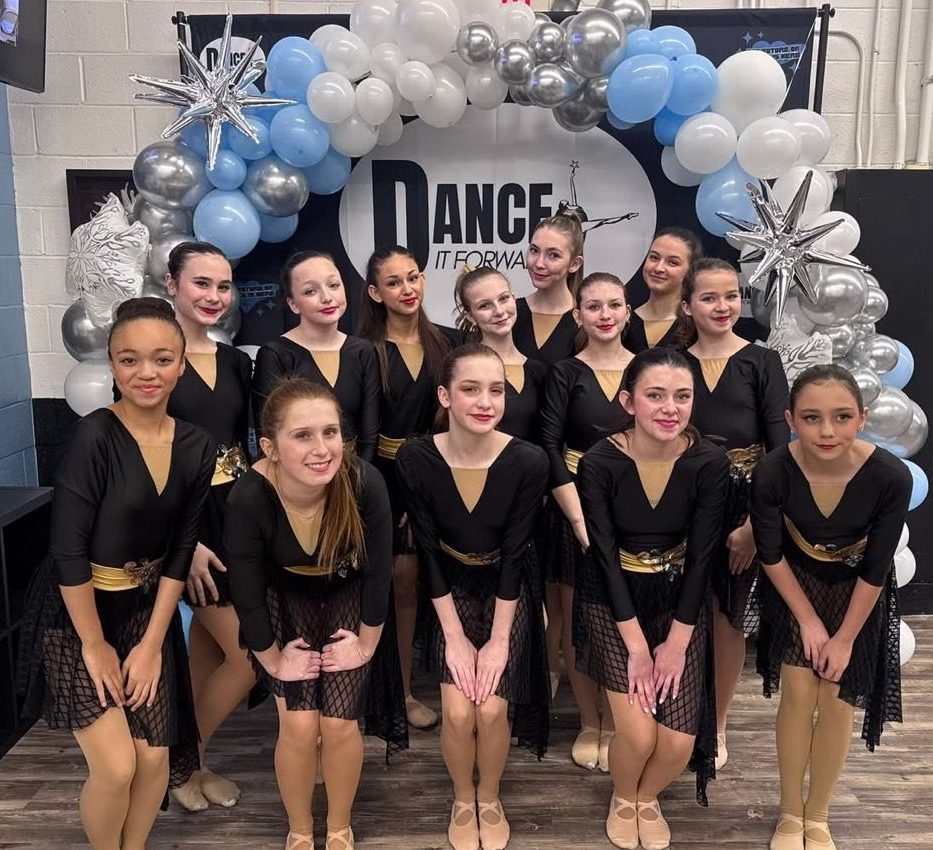 Six dancers in black outfits pose in front of a Dance competition backdrop.