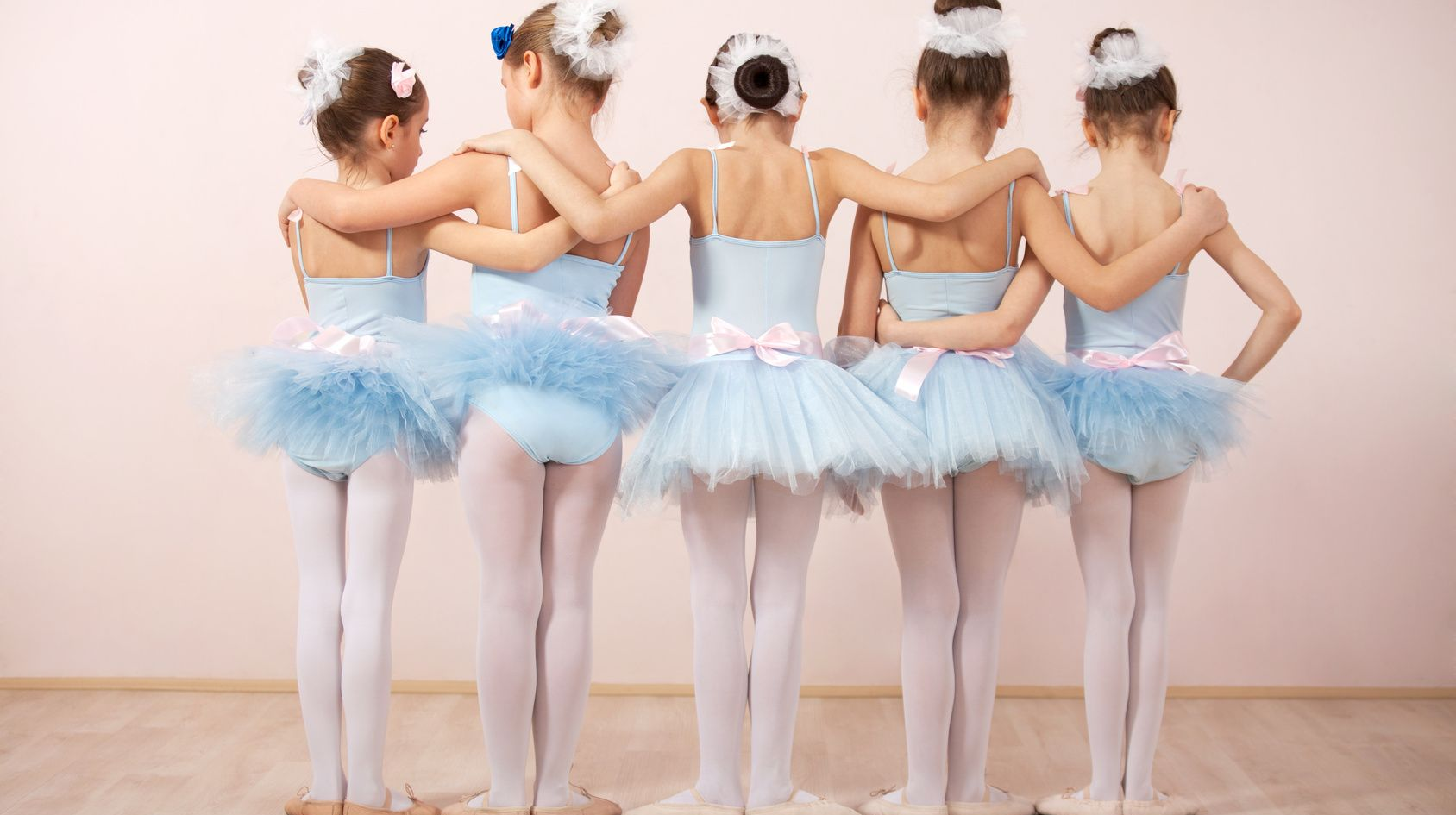 Five ballet dancers in blue tutus and white tights standing with arms around each other in a studio