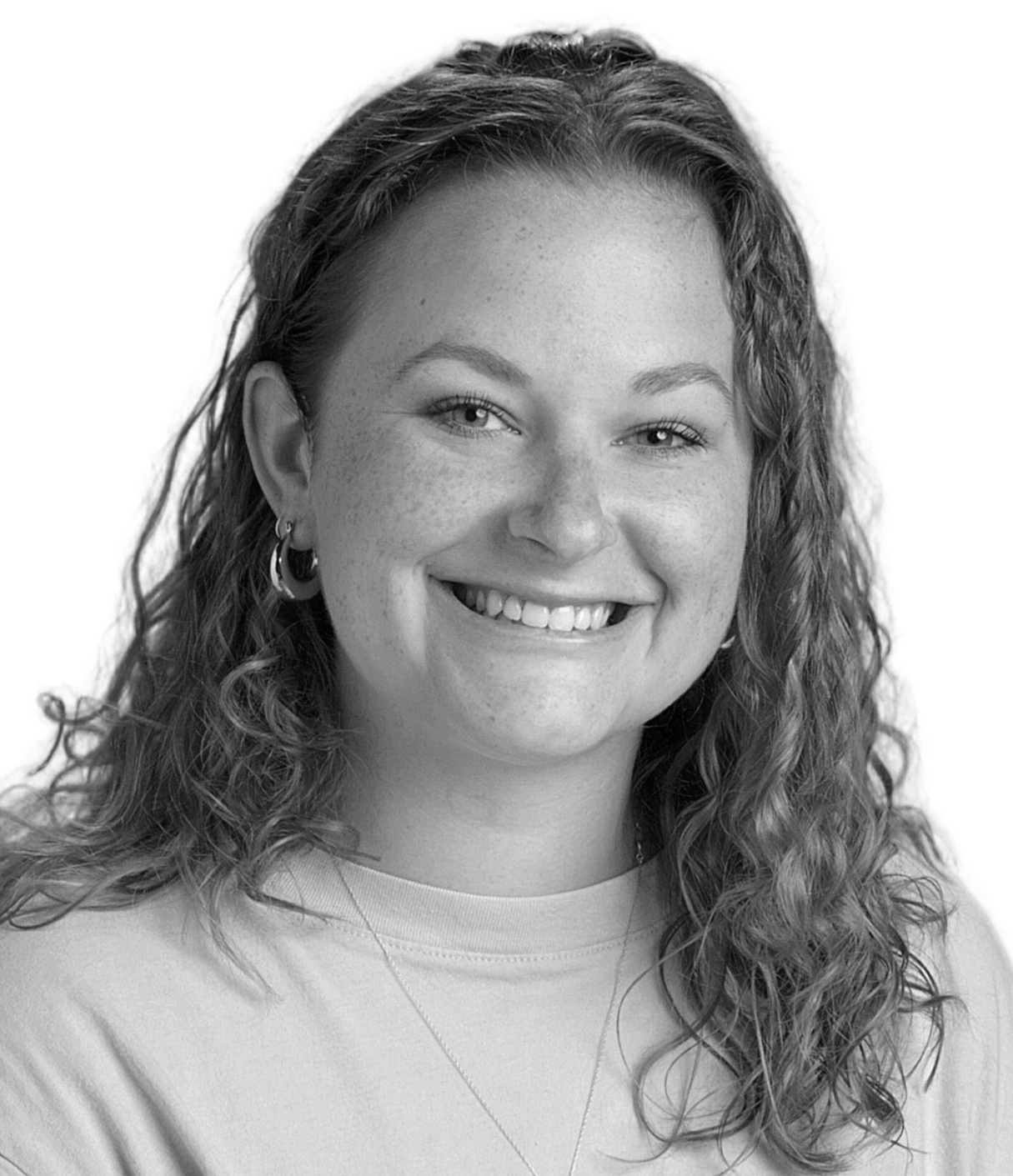 Smiling person with curly hair wearing a light shirt and necklace, facing the camera.