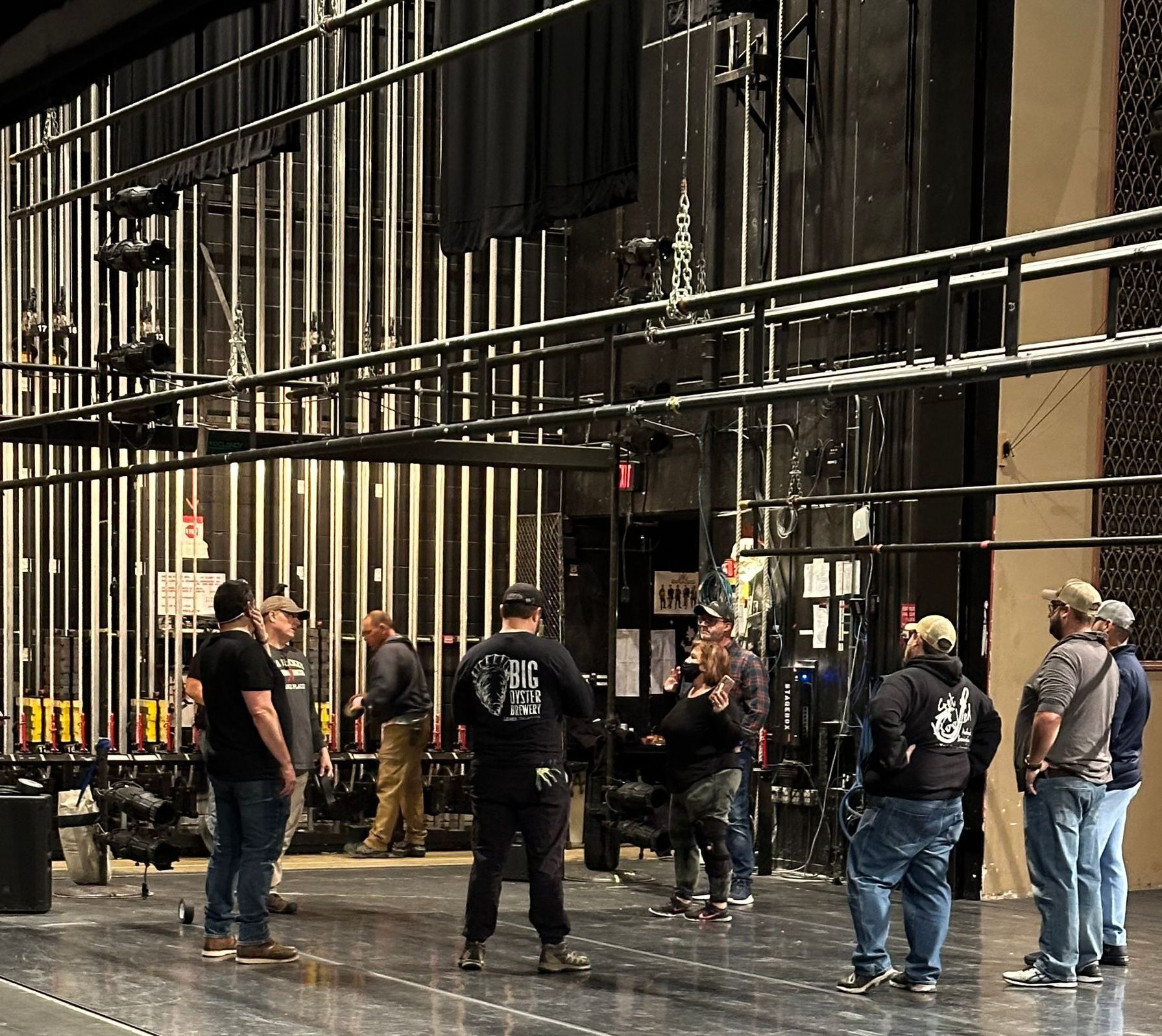 Several people stand on a theater stage beneath tall rigging and lights, facing one another in conversation.
