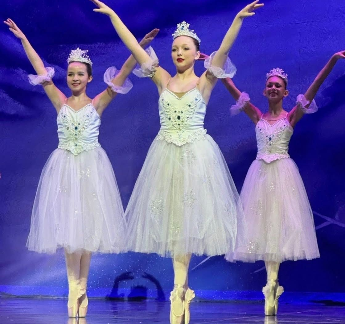 Three ballet dancers in white tutus pose on stage with arms raised under purple lighting.