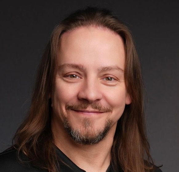Smiling person with long brown hair and a goatee against a dark background