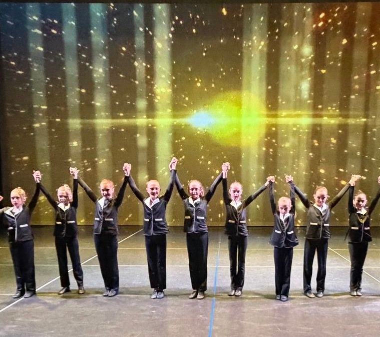 Nine performers in black outfits stand on stage with arms raised under golden lights.