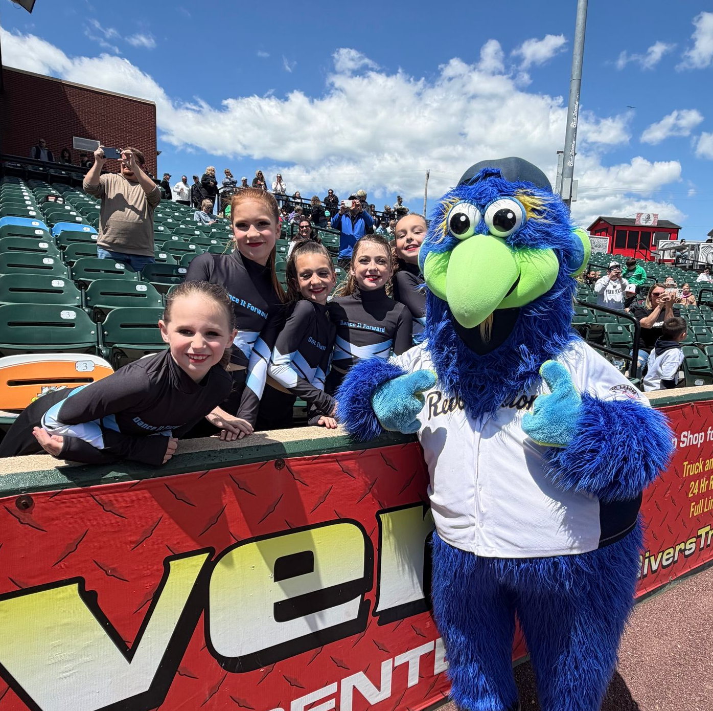 Blue mascot posing with smiling kids at a stadium barrier under a bright sky.