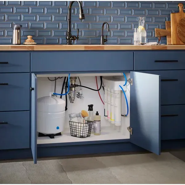 Blue kitchen cabinets with an open sink cabinet showing plumbing, a water filter, and cleaning supplies