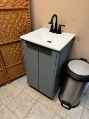 Small gray bathroom vanity with white sink and black faucet beside a stainless trash can and wicker cabinet.