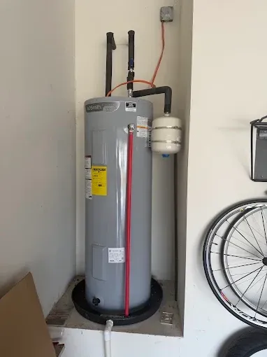 Gray water heater in a garage nook, with exposed pipes, wall outlet, and a bicycle wheel at right.
