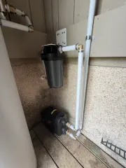 Utility room with a black filter canister and pump beside exposed pipes on a wall.