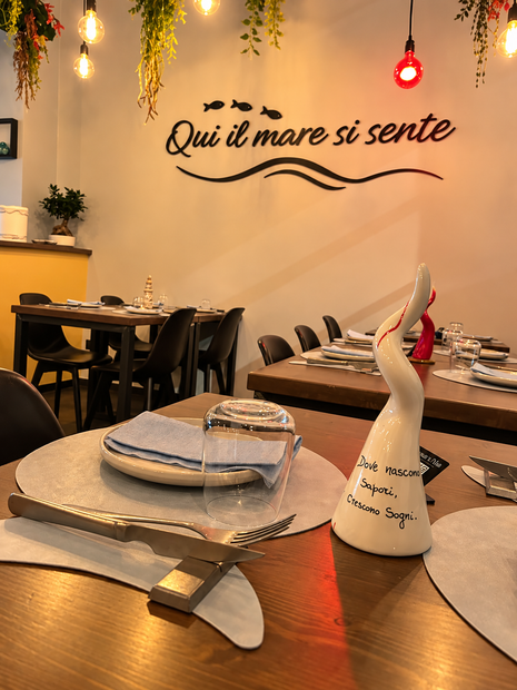 Tavolo accogliente di un ristorante con piatti, posate, una bottiglia di vino e un’insegna sulla parete con la scritta «Qui si mare si sente»