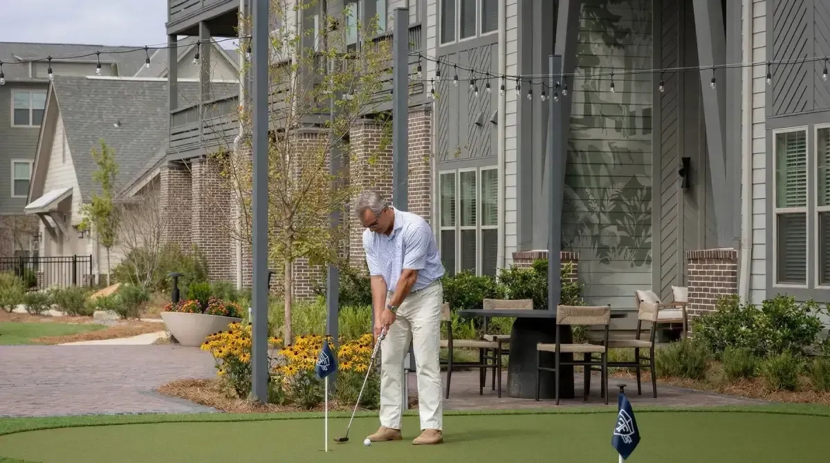 Man putts a golf ball on a green in front of apartment buildings and patio seating.