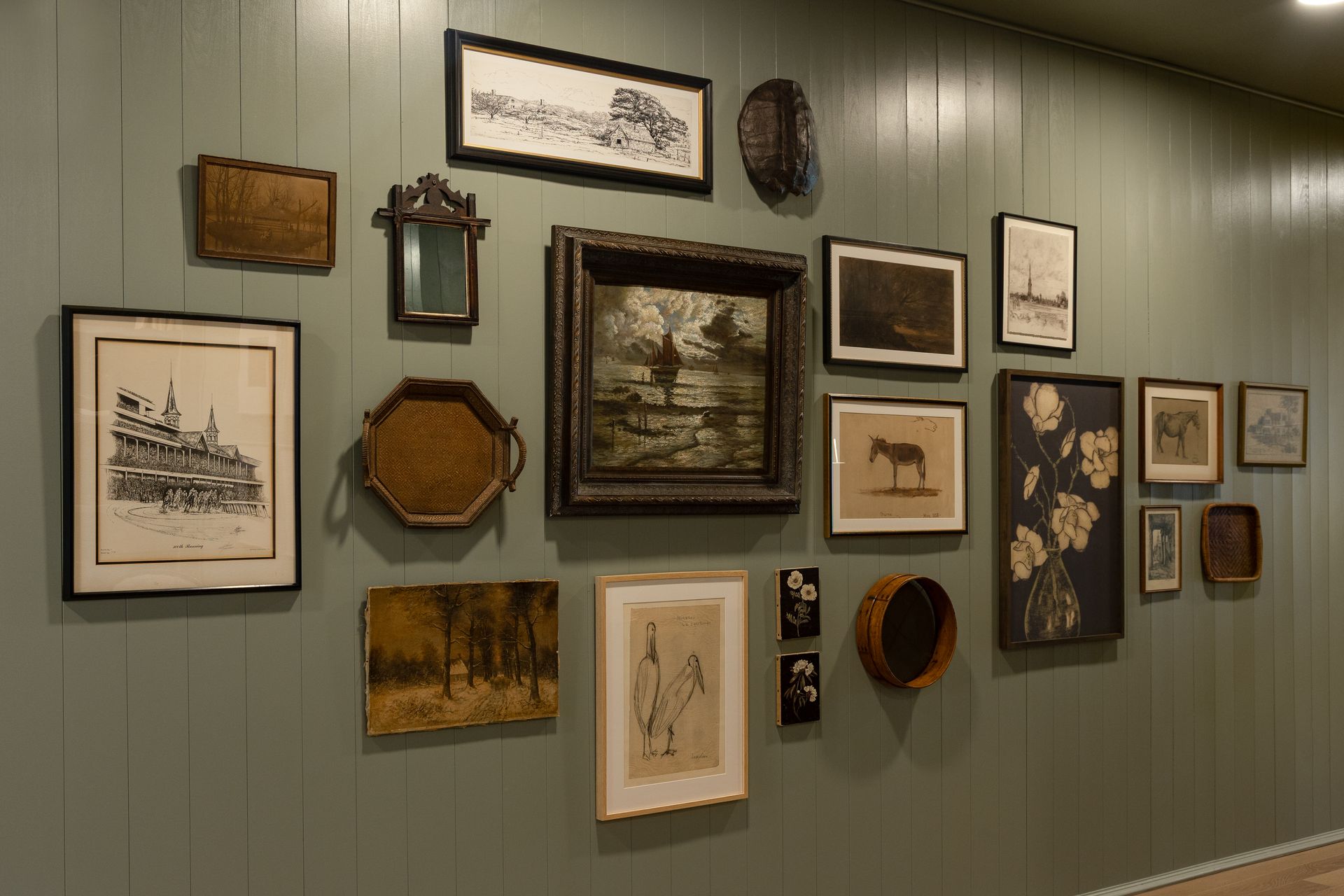 A gallery wall with various framed vintage artworks, baskets, and a small mirror, against a green paneled wall.
