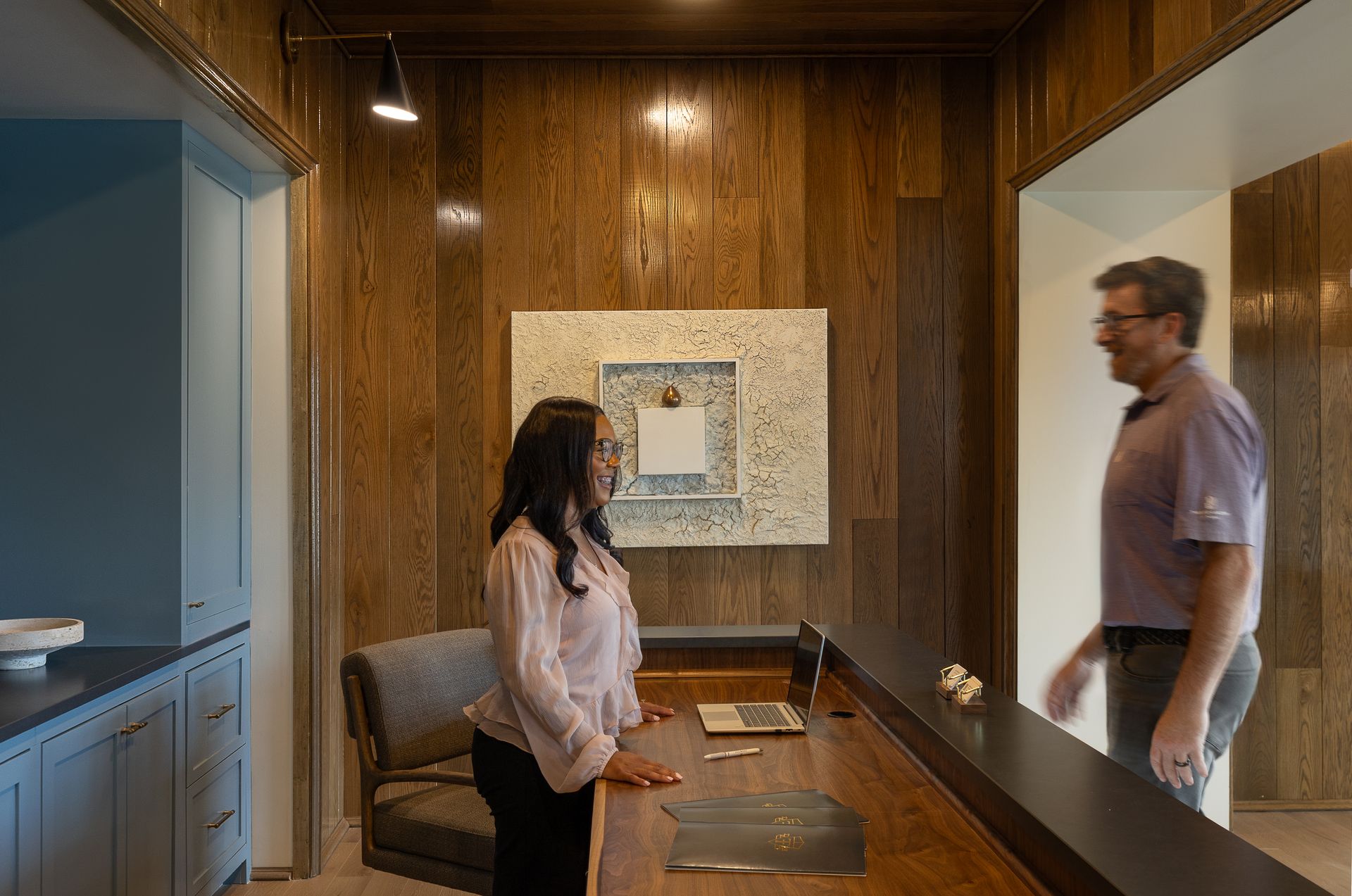 Two people standing in a wood-paneled office with a glass wall and framed art above a desk.