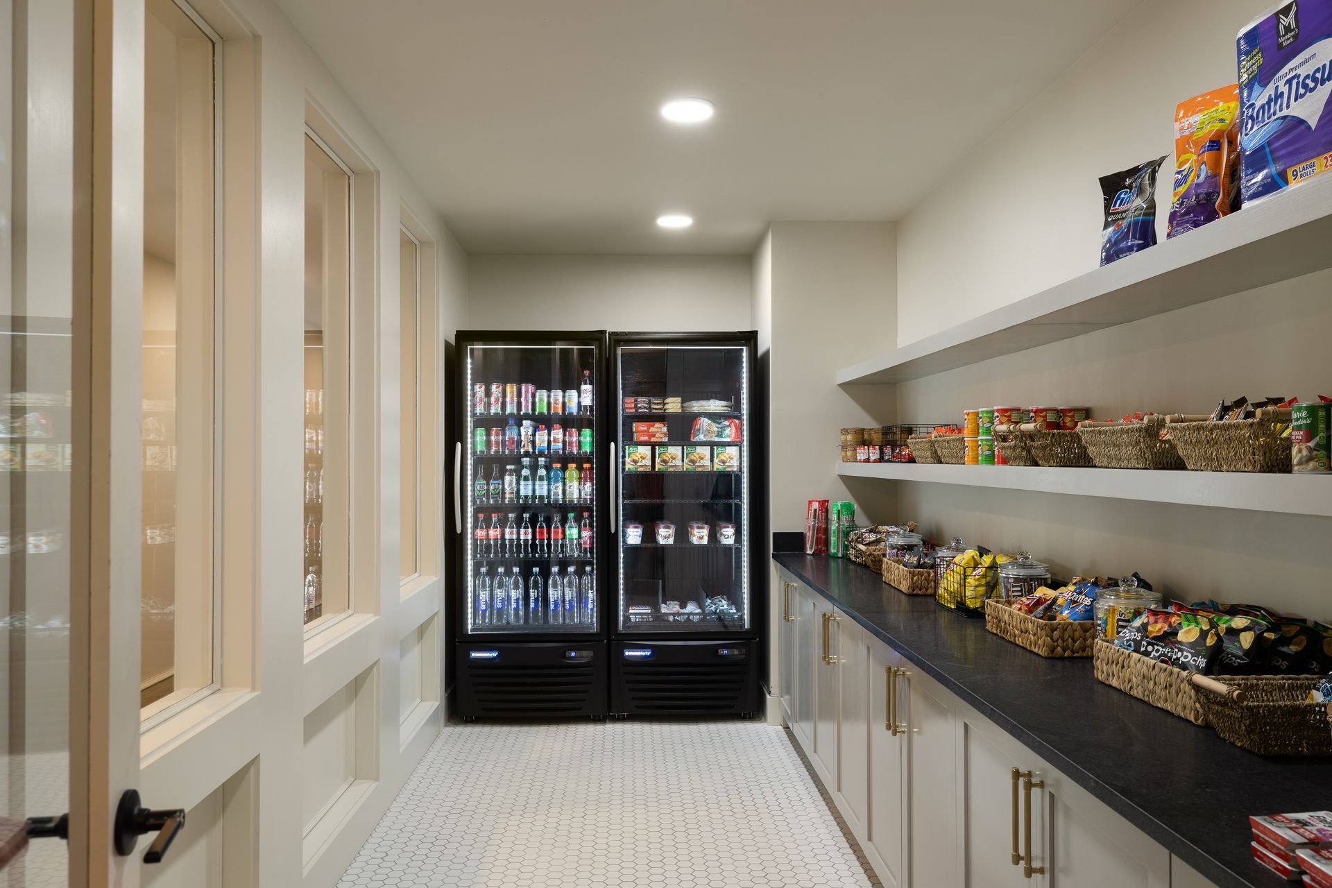 Pantry with a cooler filled with drinks, shelves stocked with snacks, and baskets. White and beige tones.