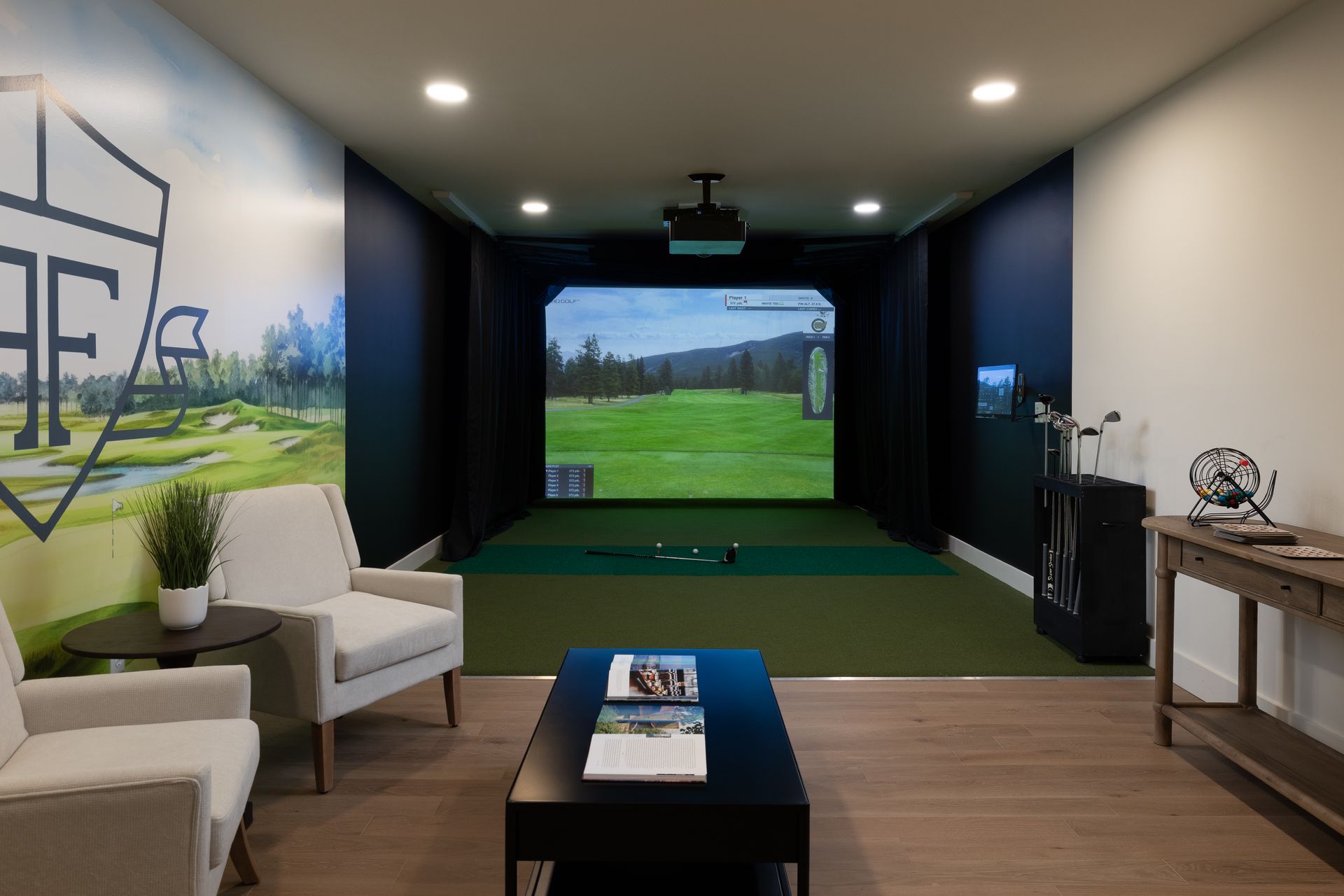 Golf simulator room with screen displaying a golf course, seating area, and golf equipment.