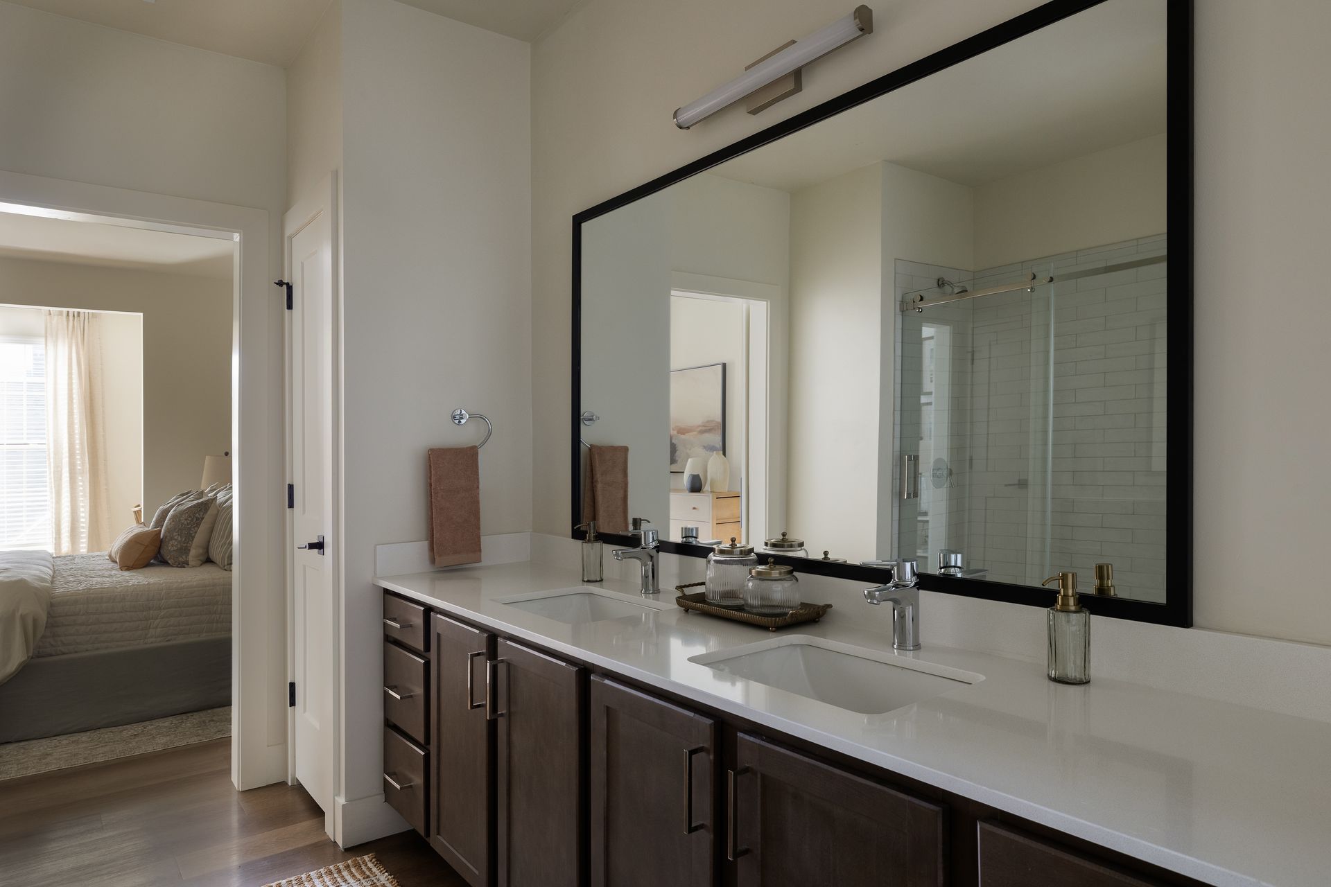 Bathroom with long mirror, double sinks, dark brown cabinets, and a bedroom in the background.