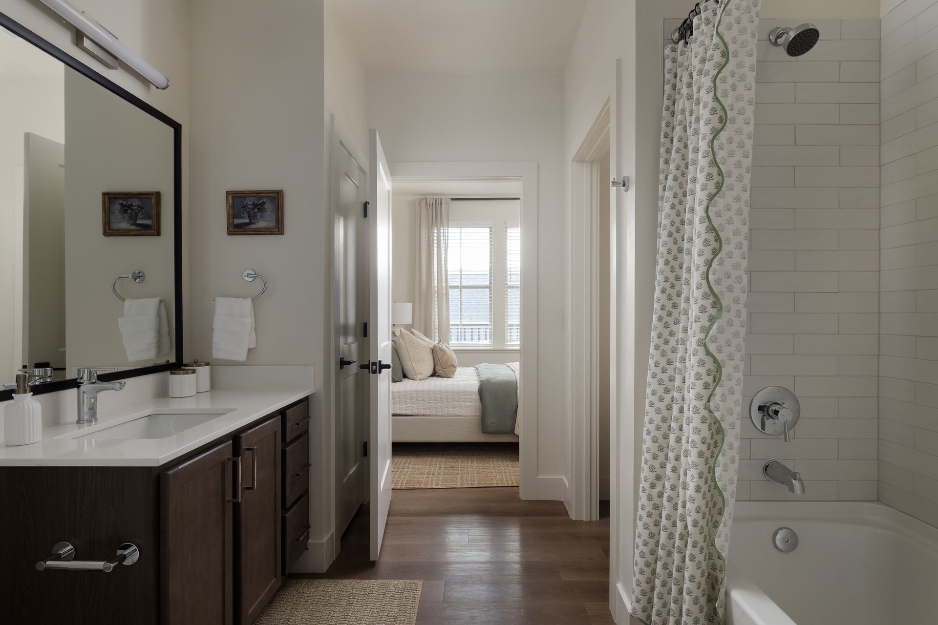 Bathroom with white walls, dark wood vanity, bathtub, and open doorway to a bedroom with bed and window.
