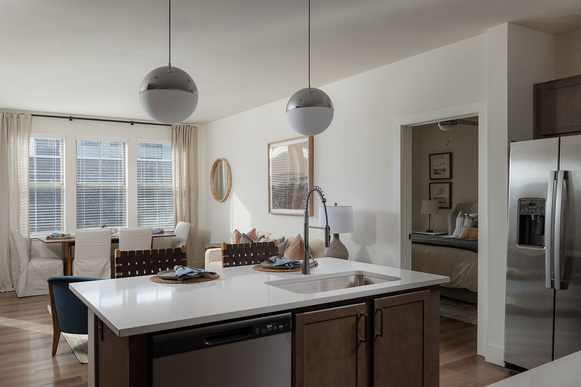 Modern kitchen with island, sink, and pendant lights. A dining area and bedroom are visible.