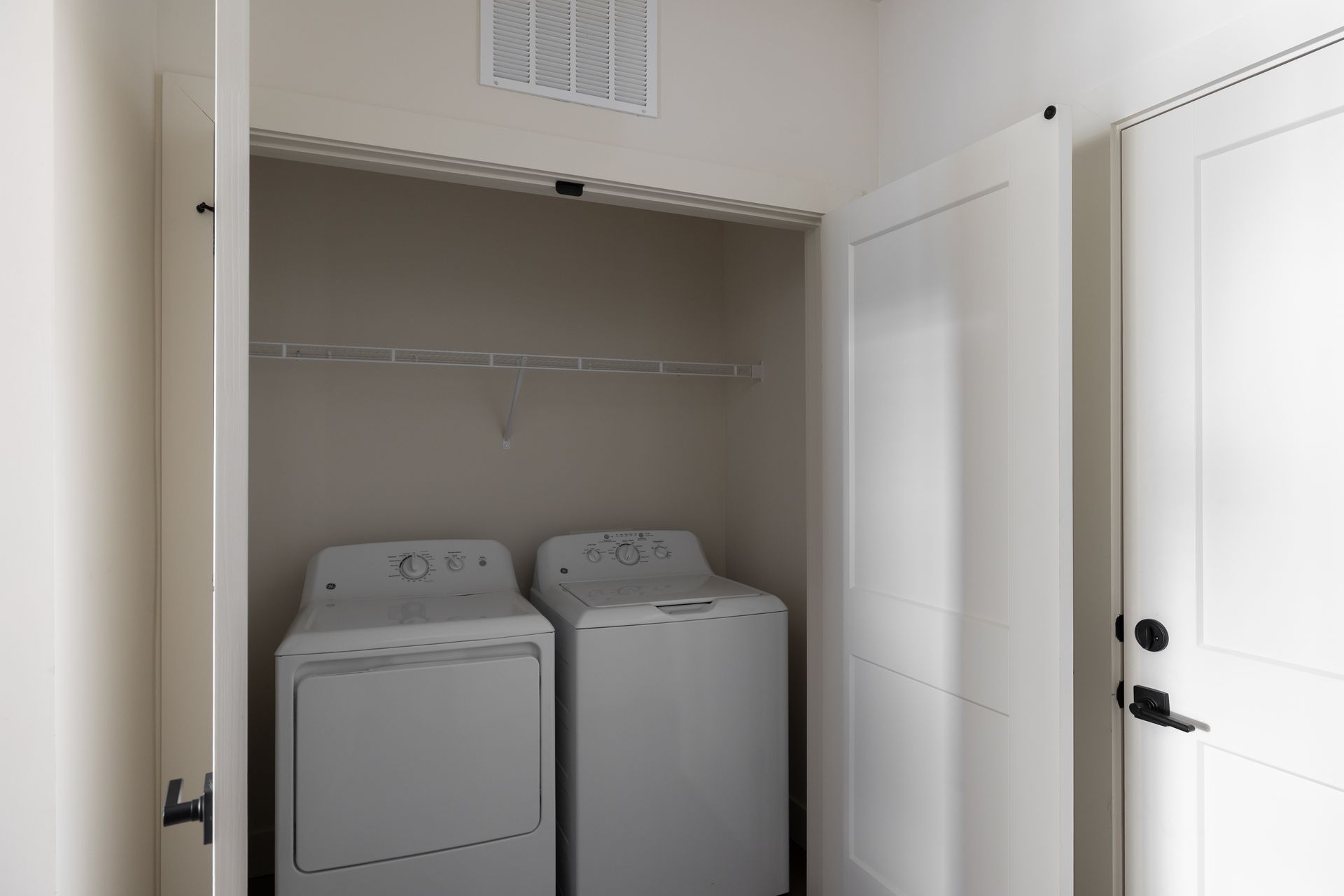 White laundry appliances inside a closet with open doors and a wire shelf above.
