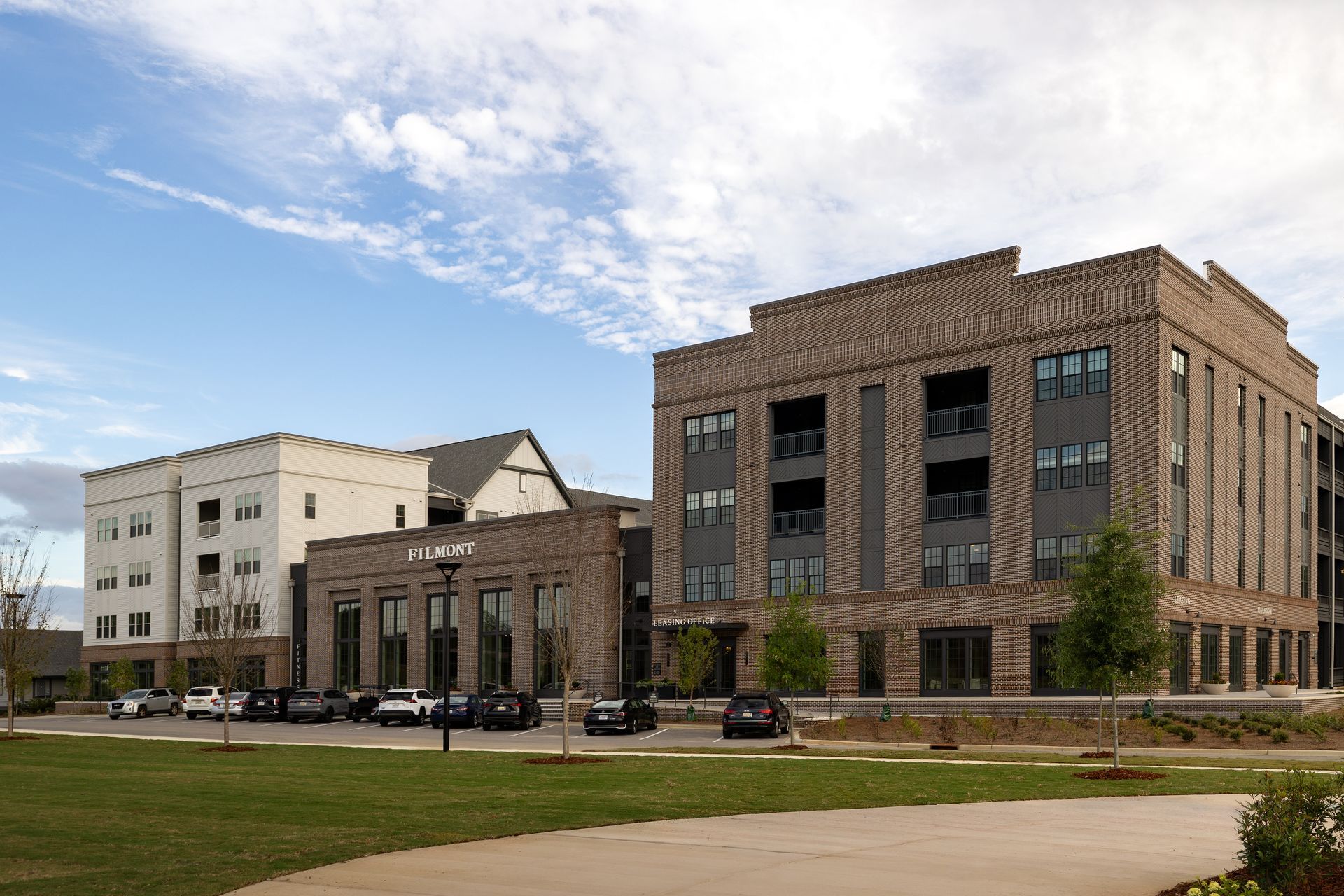 Modern brick and white multi-story apartment buildings on a grassy field, under a cloudy blue sky.