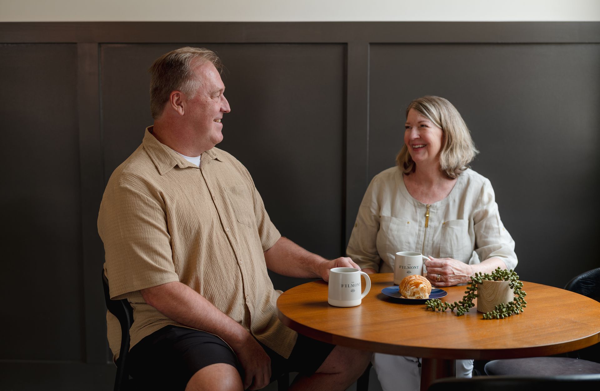 Two people smiling and talking at a small round table with mugs and snacks in a cozy room.