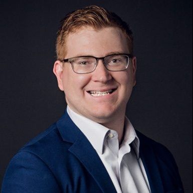 Smiling man in glasses wearing a blue suit jacket and white shirt against a dark background