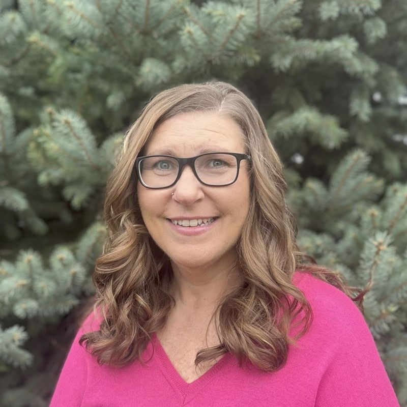 Woman with glasses smiling outdoors in a bright pink top, standing in front of evergreen trees