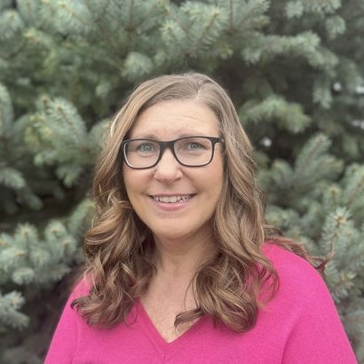Woman with glasses smiling outdoors in a bright pink top, standing in front of evergreen trees