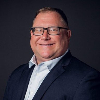 Portrait of a smiling man in glasses wearing a navy suit and light blue shirt on a dark background