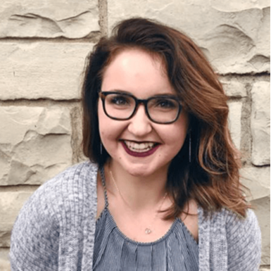 Smiling woman with glasses and shoulder-length hair, wearing a gray cardigan against a stone wall