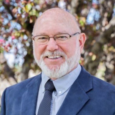 Smiling bald man in glasses wearing a blue suit and tie outdoors, with blurred trees behind him