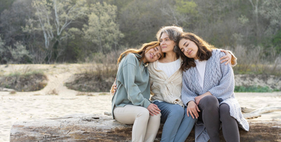 Three friends sit close together on a log outdoors, smiling in a natural landscape.