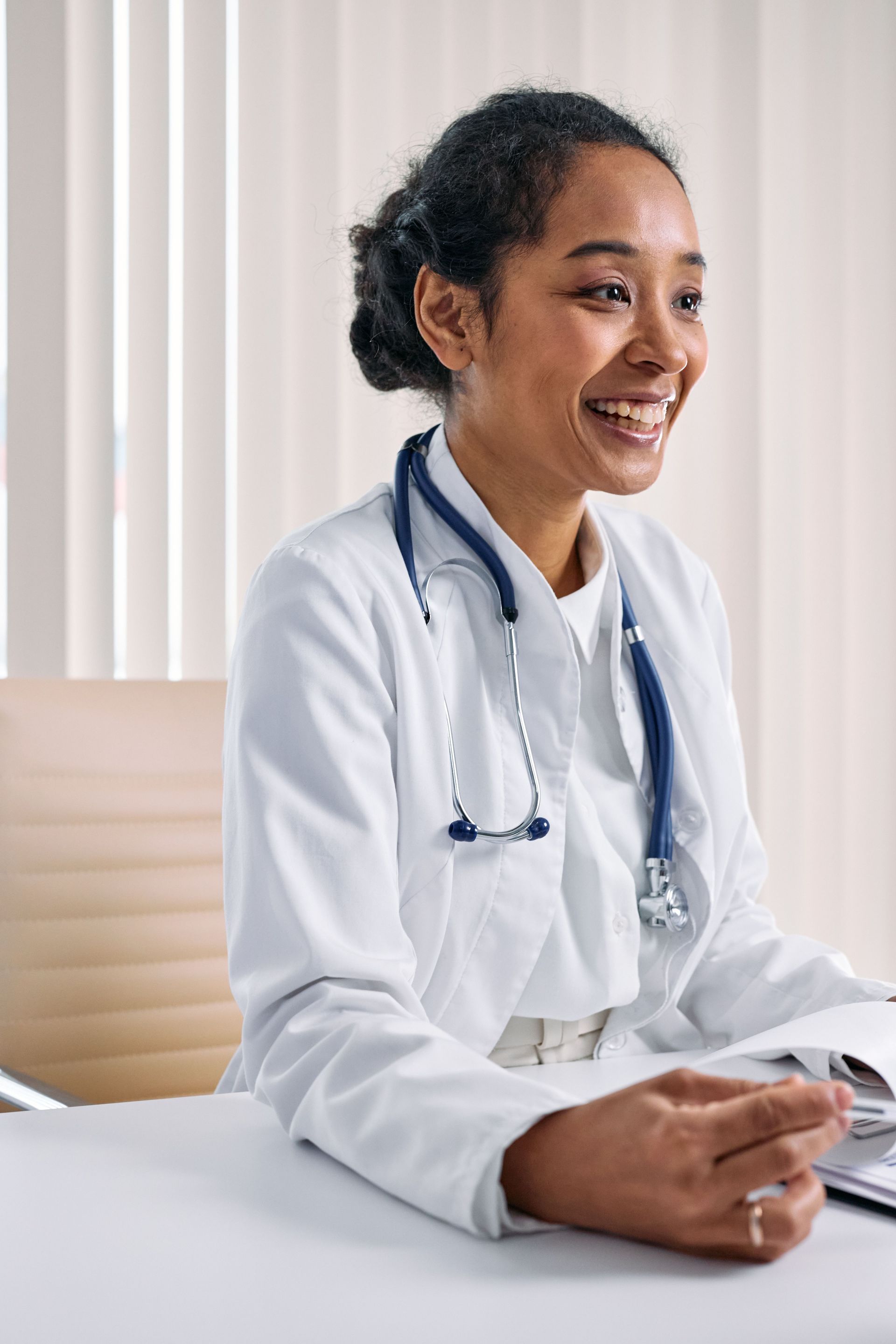 Doctor in a white coat smiling at a desk with a stethoscope around her neck
